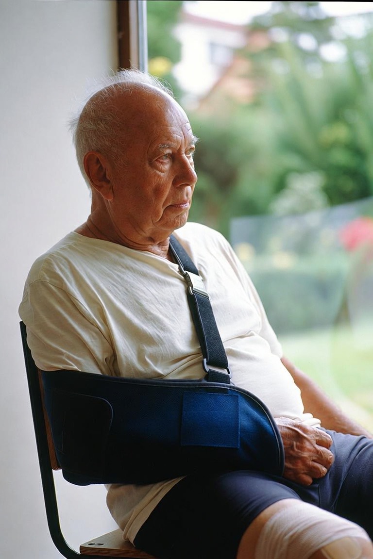 An older man in a shoulder sling sitting by a window at home, looking out at the garden.