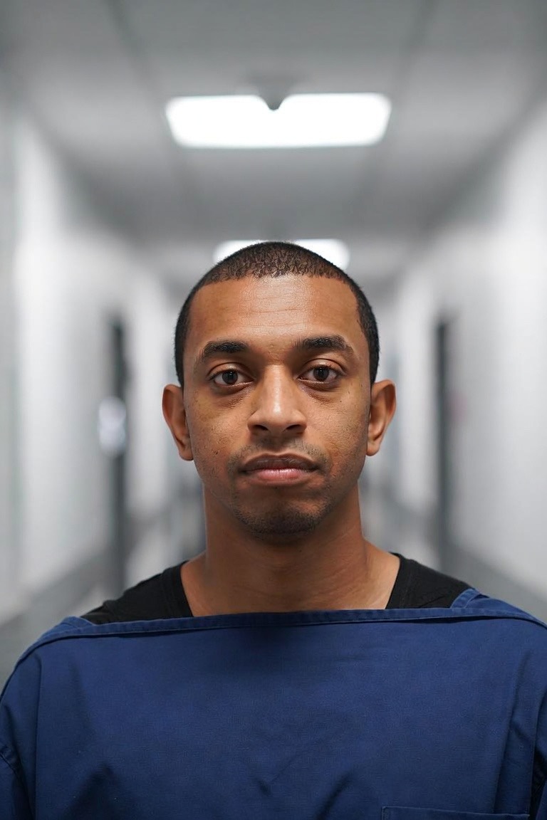 A physiotherapist in scrubs looking straight into the camera, in the same corridor and at the same distance as the patient portrait.